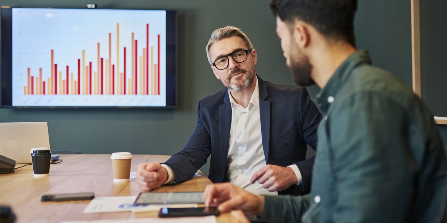 Two business professionals, one older and one younger, are sitting in a conference room having a serious discussion. They are both looking at the younger man's tablet. The older man has a thoughtful expression on his face, while the younger man is looking at him intently.