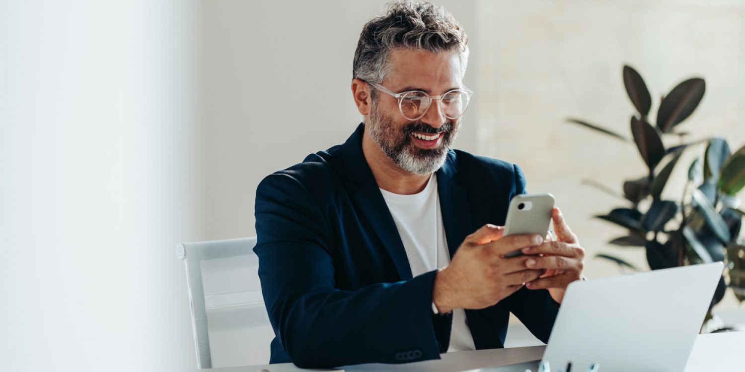 Cheerful, mature professional man is engrossed in his smartphone while sitting at a white desk in a sunny, contemporary office space. His laptop and notepad are at hand, suggesting productivity and connectivity.