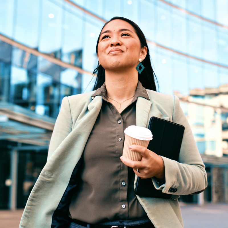 Smiling businesswoman holding coffee and documents while walking outside a modern office building. Corporate environment, confidence, and urban lifestyle.