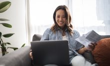 An Asian woman sits relaxed on a sofa, using a laptop and holding documents, smiling as she manages her finances and enjoys working from home.