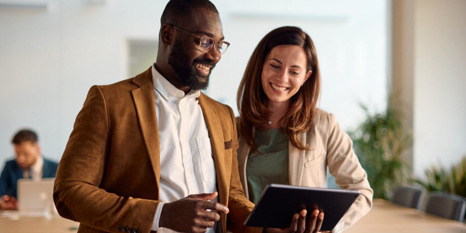 Two happy business colleagues engaging with a digital tablet and discussing various projects while standing in a bright, modern office environment, showcasing teamwork and collaboration