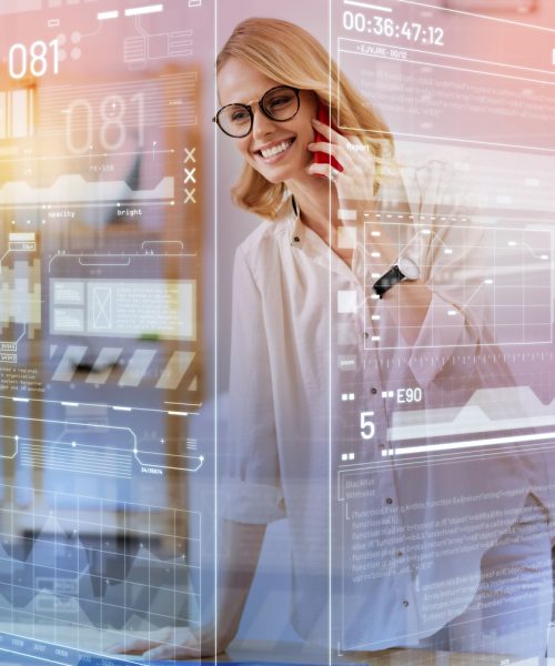 Phone talk. Optimistic skilled experienced engineer standing next to the table with a big blueprint on it and smiling while talking on the phone with her cheerful colleague