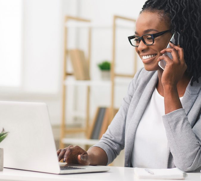 Female Entrepreneur. Cheerful African American Businesswoman Talking On Phone Working On Laptop In Modern Office. Empty Space